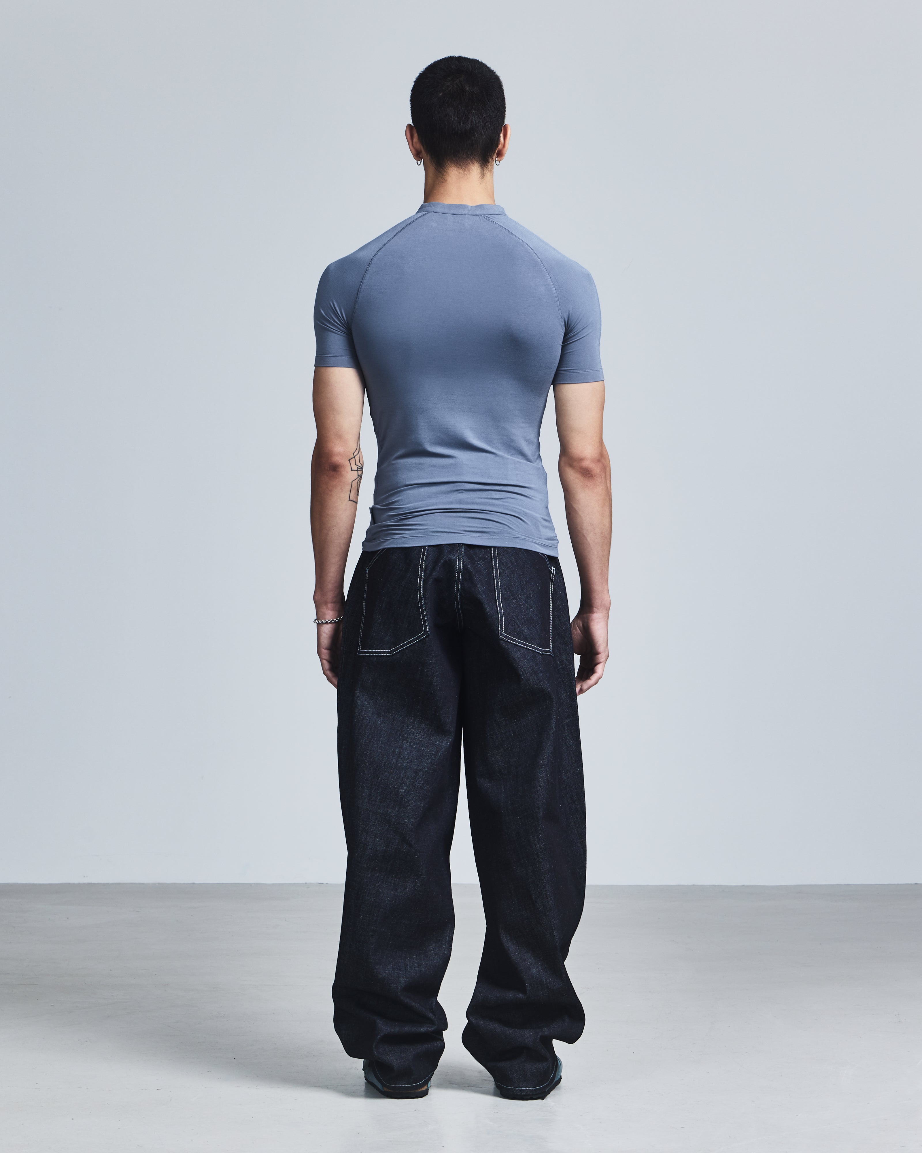 Man wearing a blue t-shirt and dark jeans standing against a light gray background